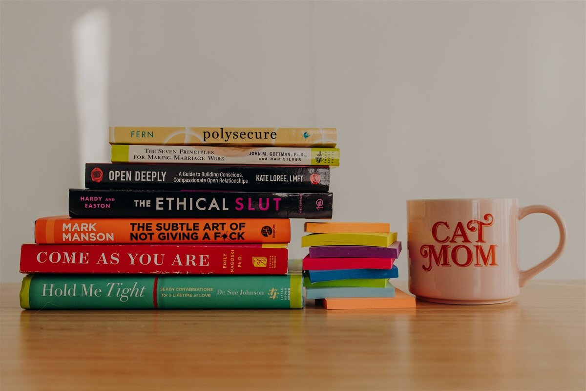 stack of sex books on desk next to sticky notes and a cat mom mug