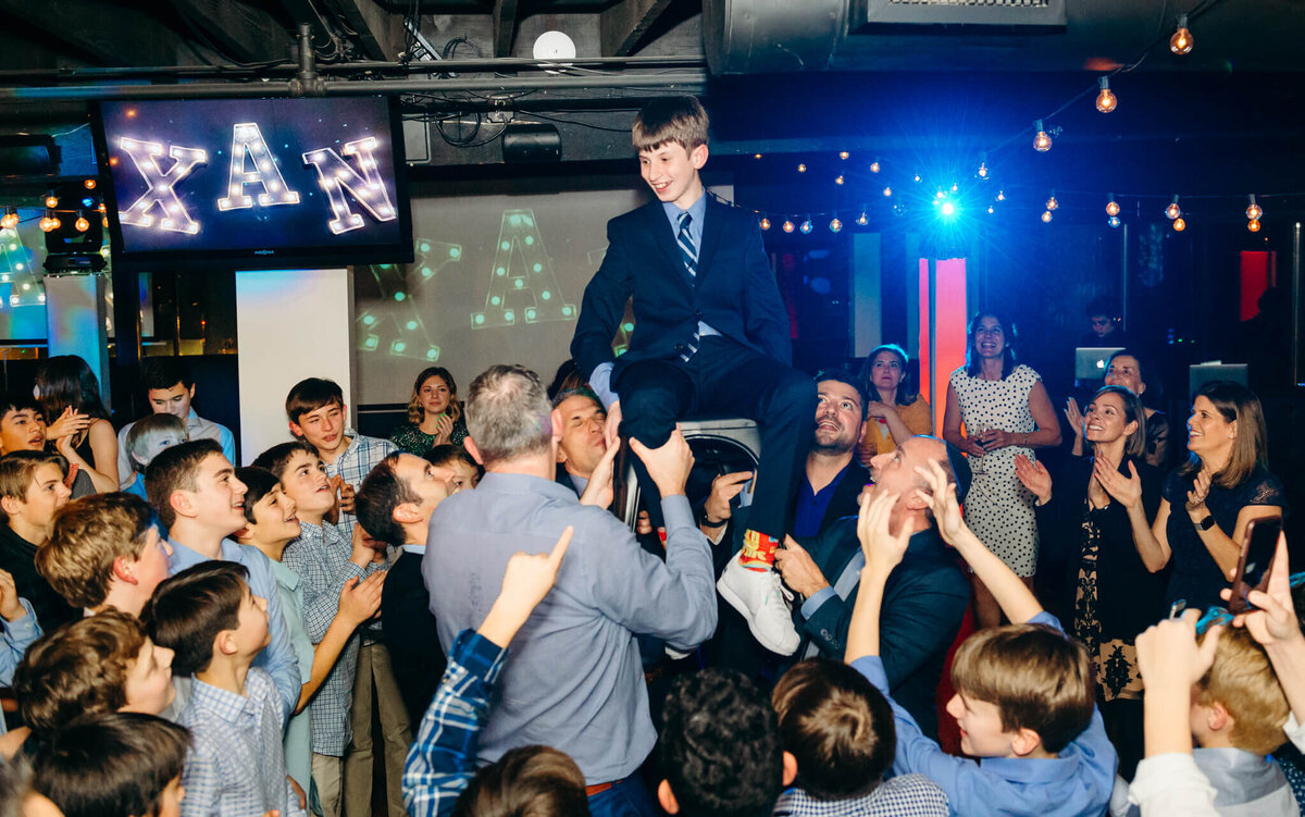 A crowd celebrates a boy for his mitzvah on the dance floor lifting him in the air in a chair