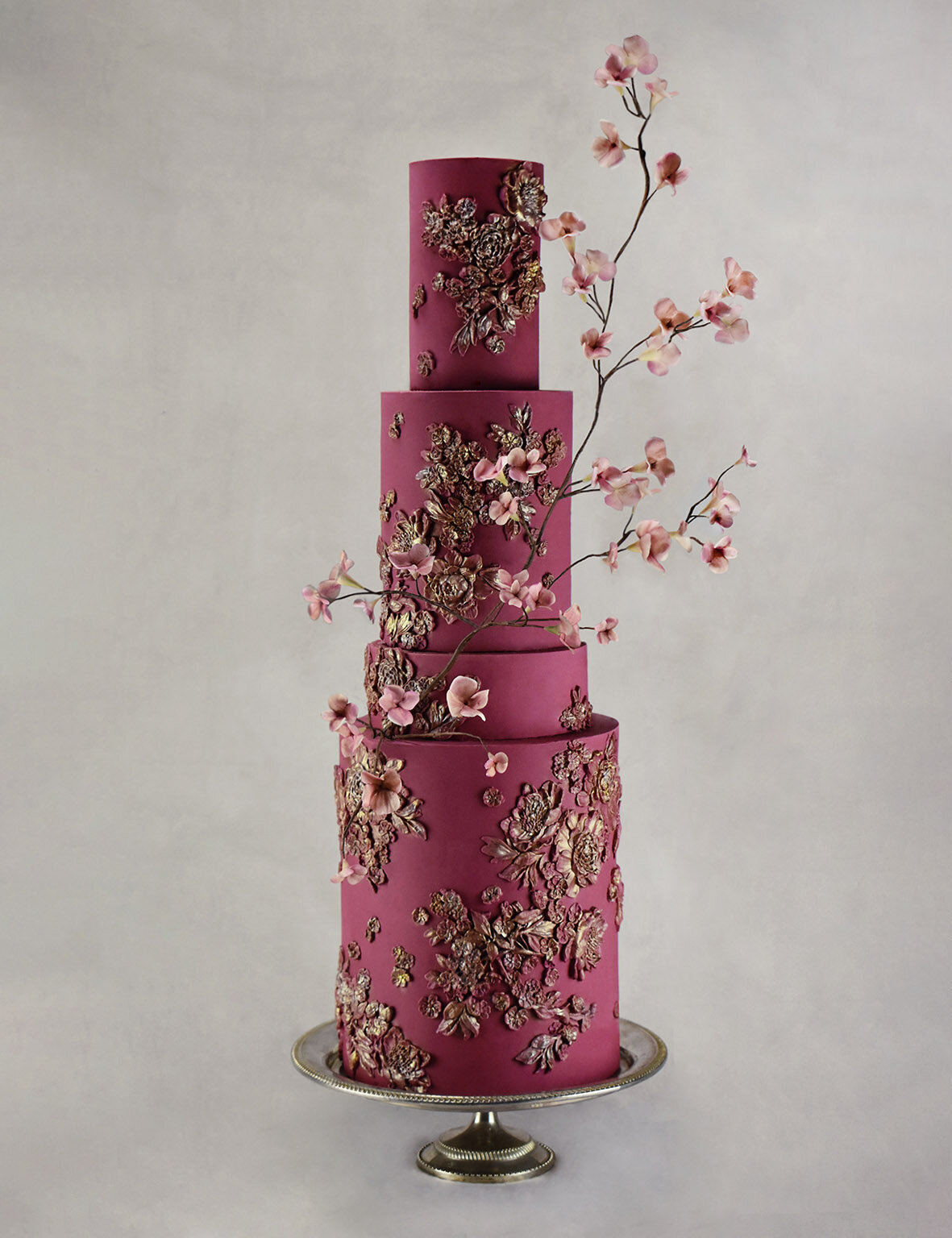 A large four tiered burgundy  wedding cake with floral bas relief and a sugar blossom branch