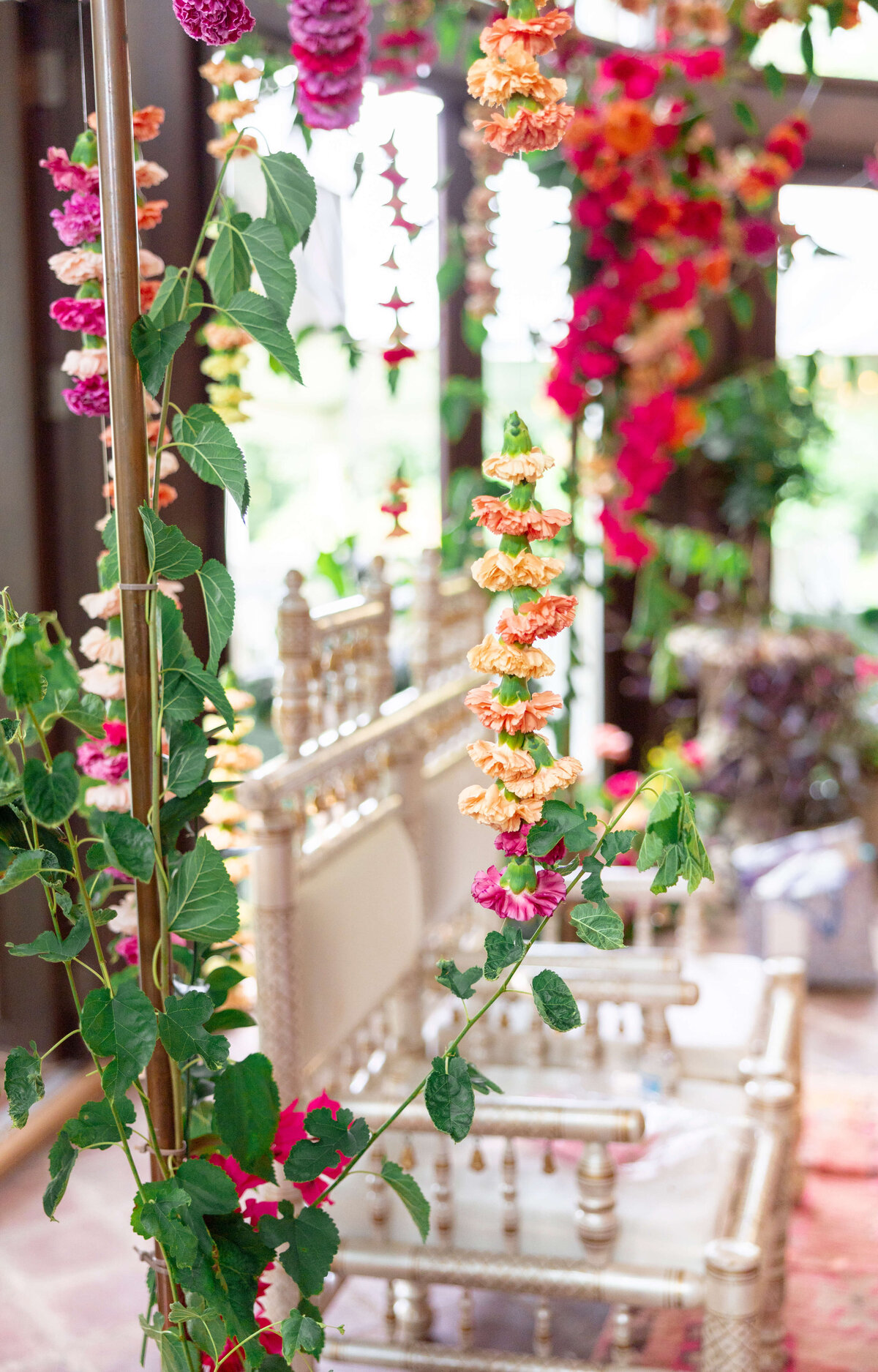 A decorative scene featuring ornate wooden chairs and vibrant floral garlands in pink, orange, and yellow, draped across a metal rod. The setting appears colorful and festive, with soft natural light filtering in.