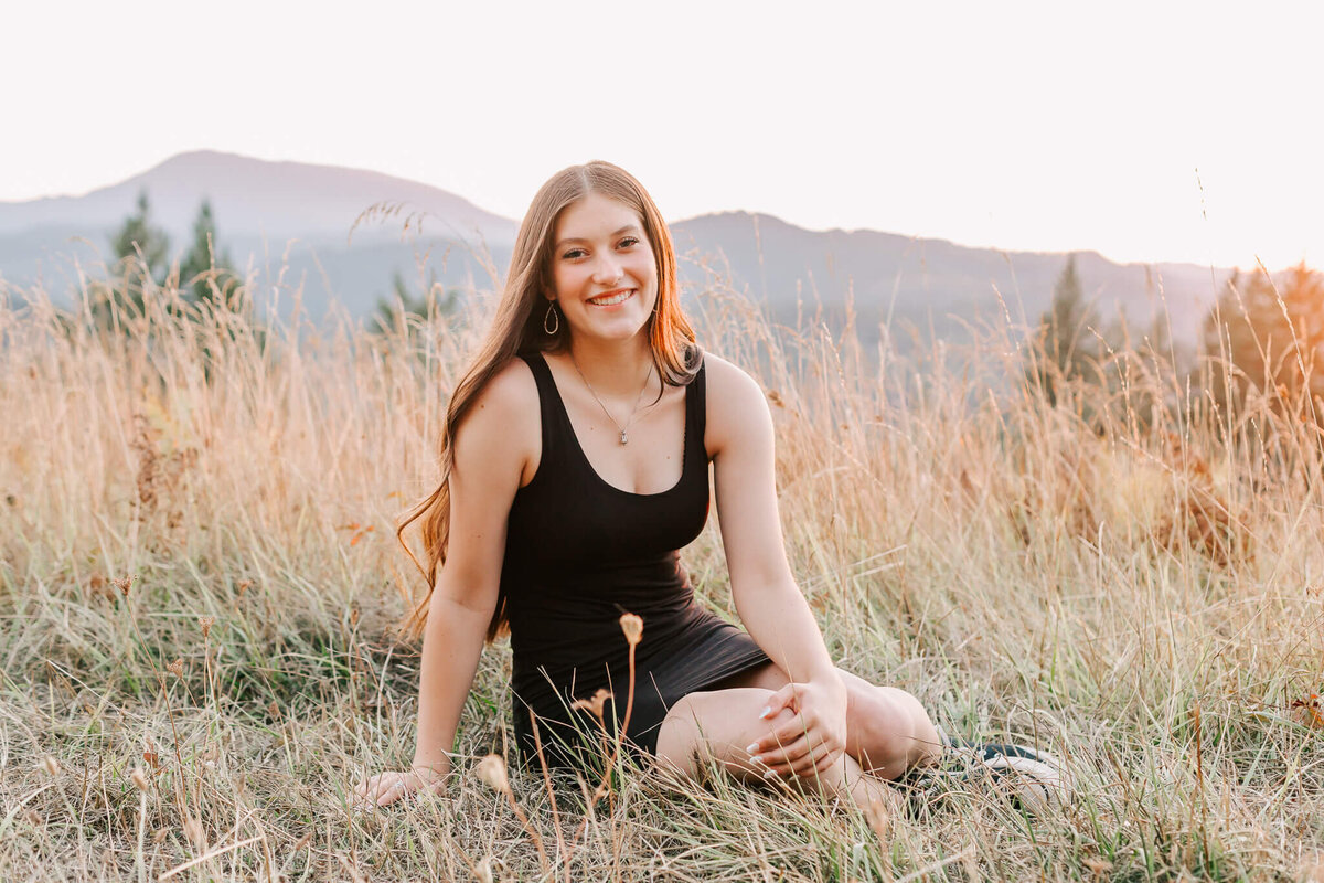 A young woman sits in the grass smiling with the mountains behind her