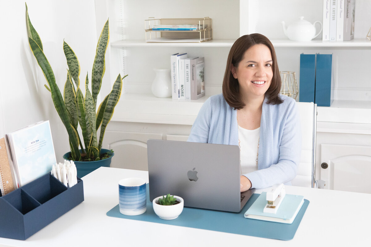 professional young woman in a blue cardigan sits at a white desk with a laptop in a bright office with a green plant in the background New York branding photographer Milestone Images