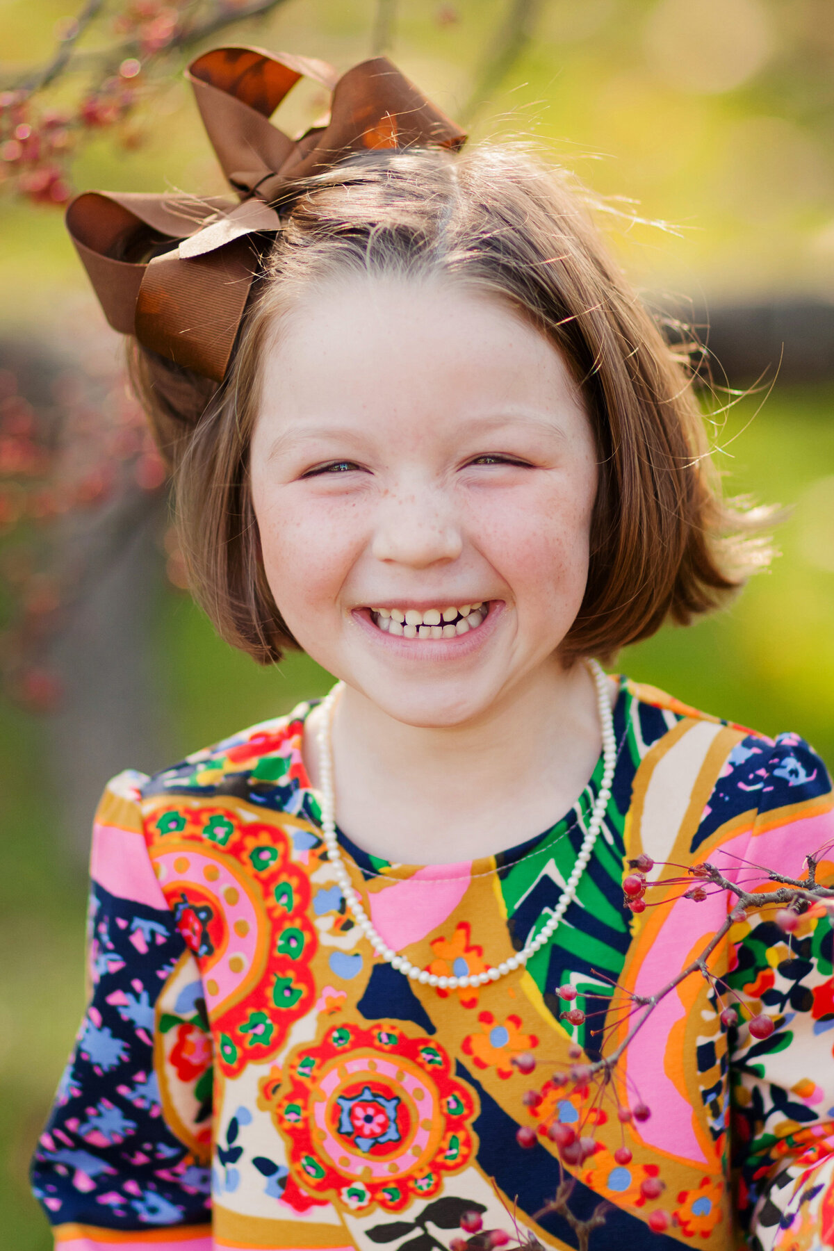 Girl with a brightly colored top, pearl necklace, and big brown bow in her hair smiles big at the camera with happy eyes.