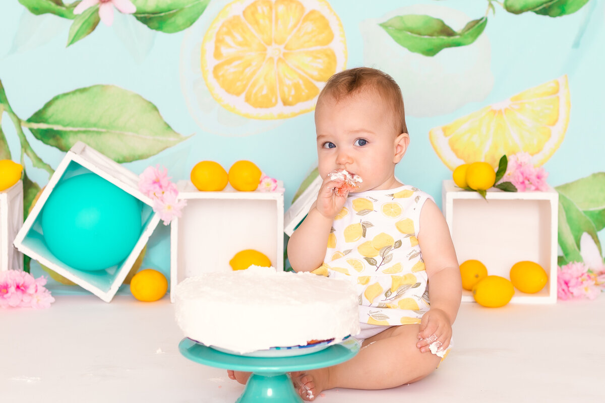 Cake Smash Photographer, a baby eats cake wears a onesie with yellow flowers and sits before a backdrop with lemons