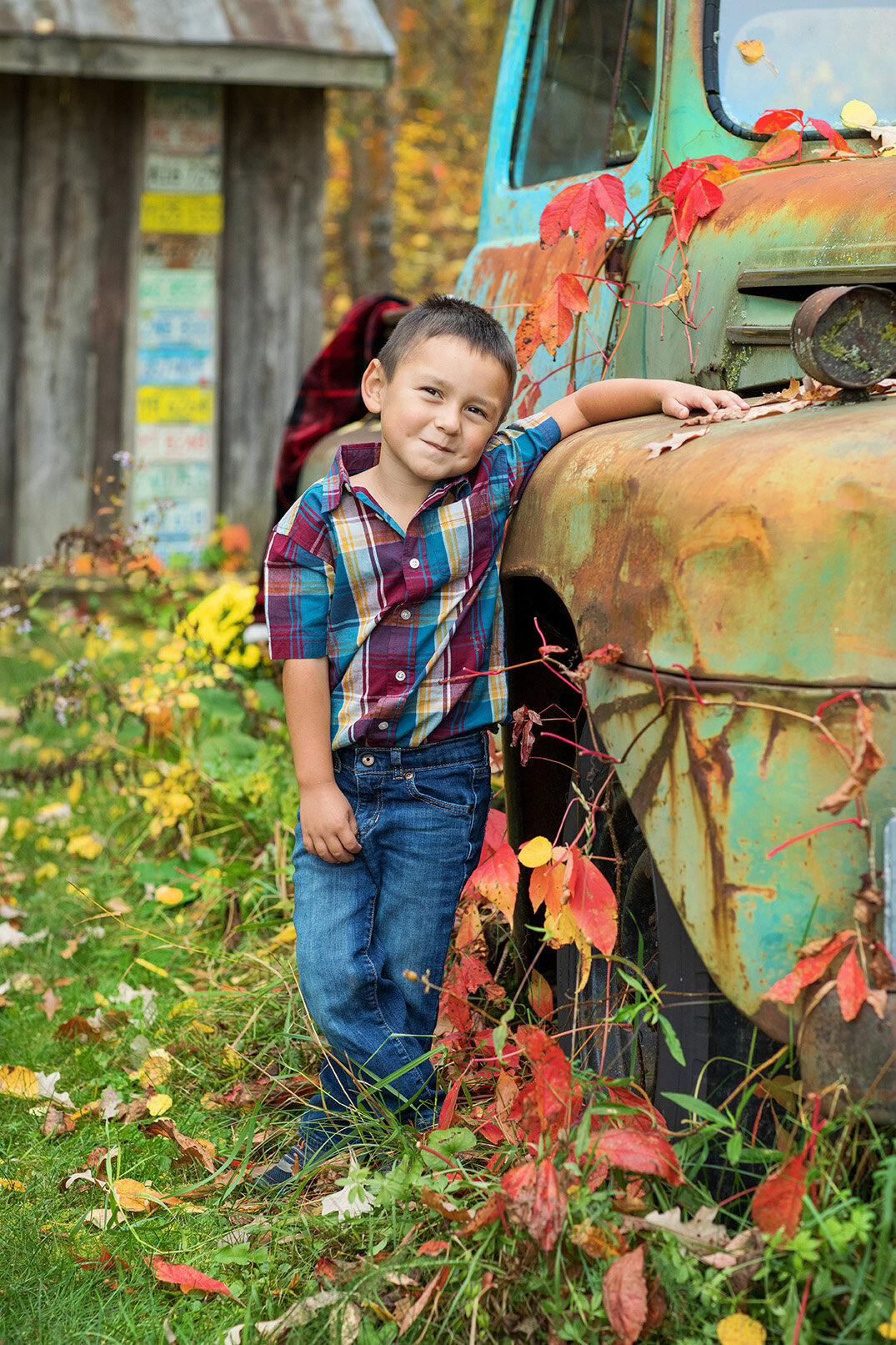 Young boy standing next to an old Ford pickup with his hand on the truck with Fall colored leaves on a vine growing up the truck.