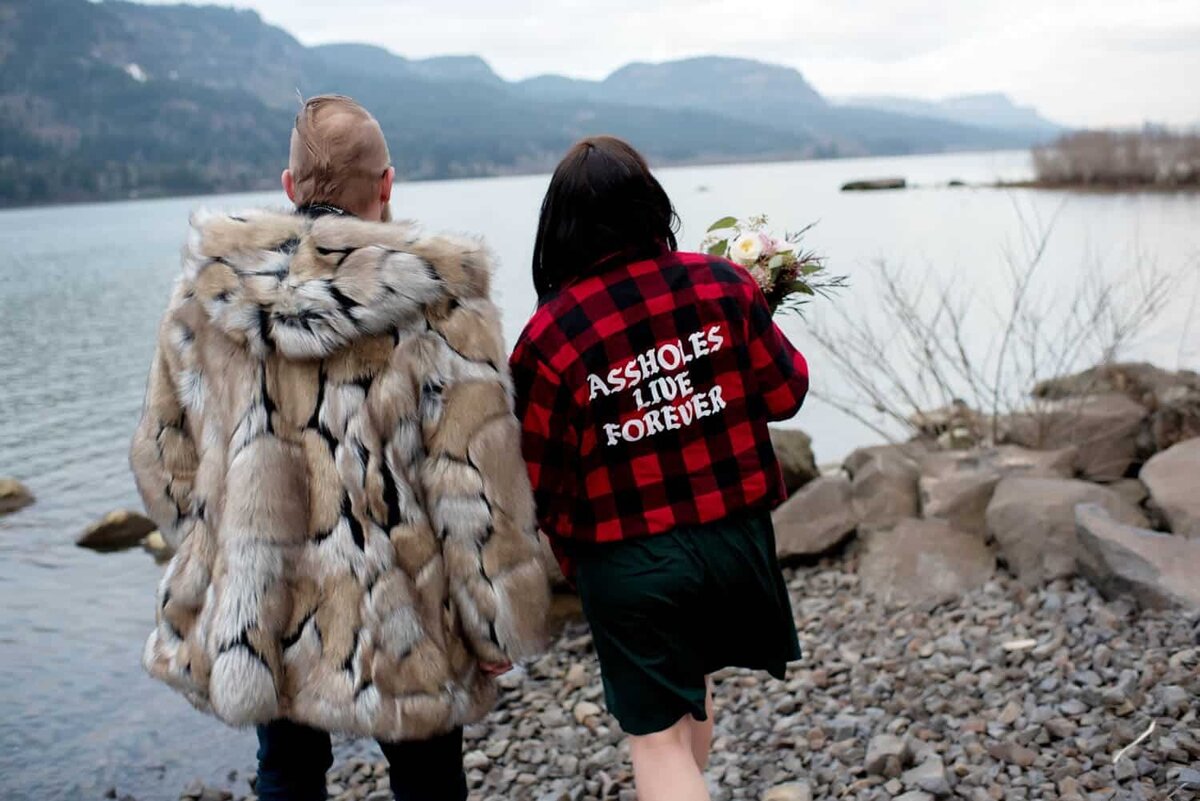a man in a fun faux fur coat and a woman wearing a plaid jacket that says "assholes live forever" hold hands while looking out at the columbia river gorge at dalton point