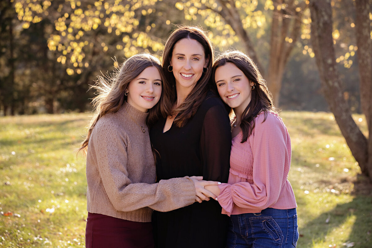 Happy mother with HS senior and middle school daughter standing together in green field with sloping hill and tree at Anne Springs Close Greenway Charlotte NC