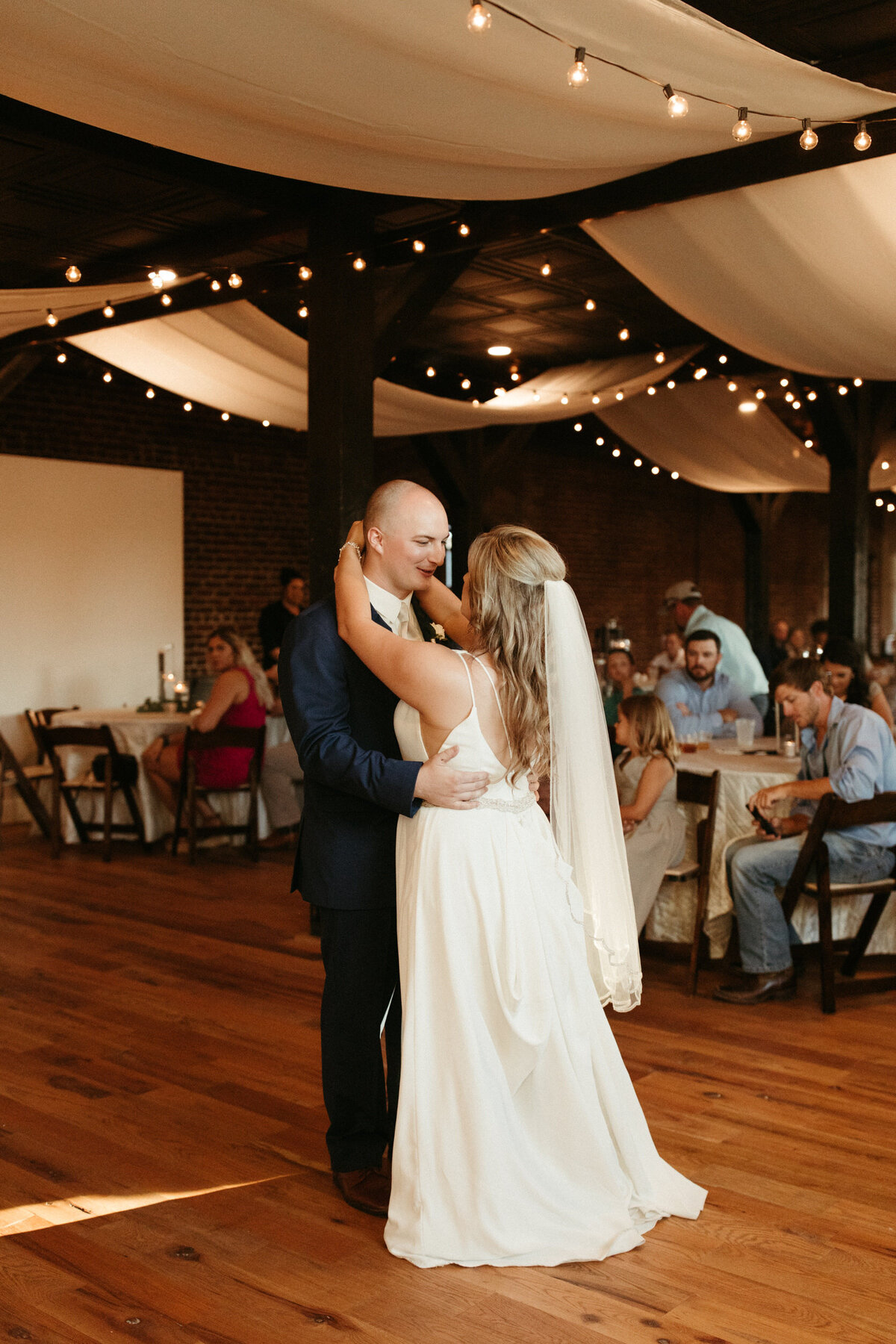 Bride and groom dancing during their first dance in their wedding reception hall with string lights and drapery hanging above them