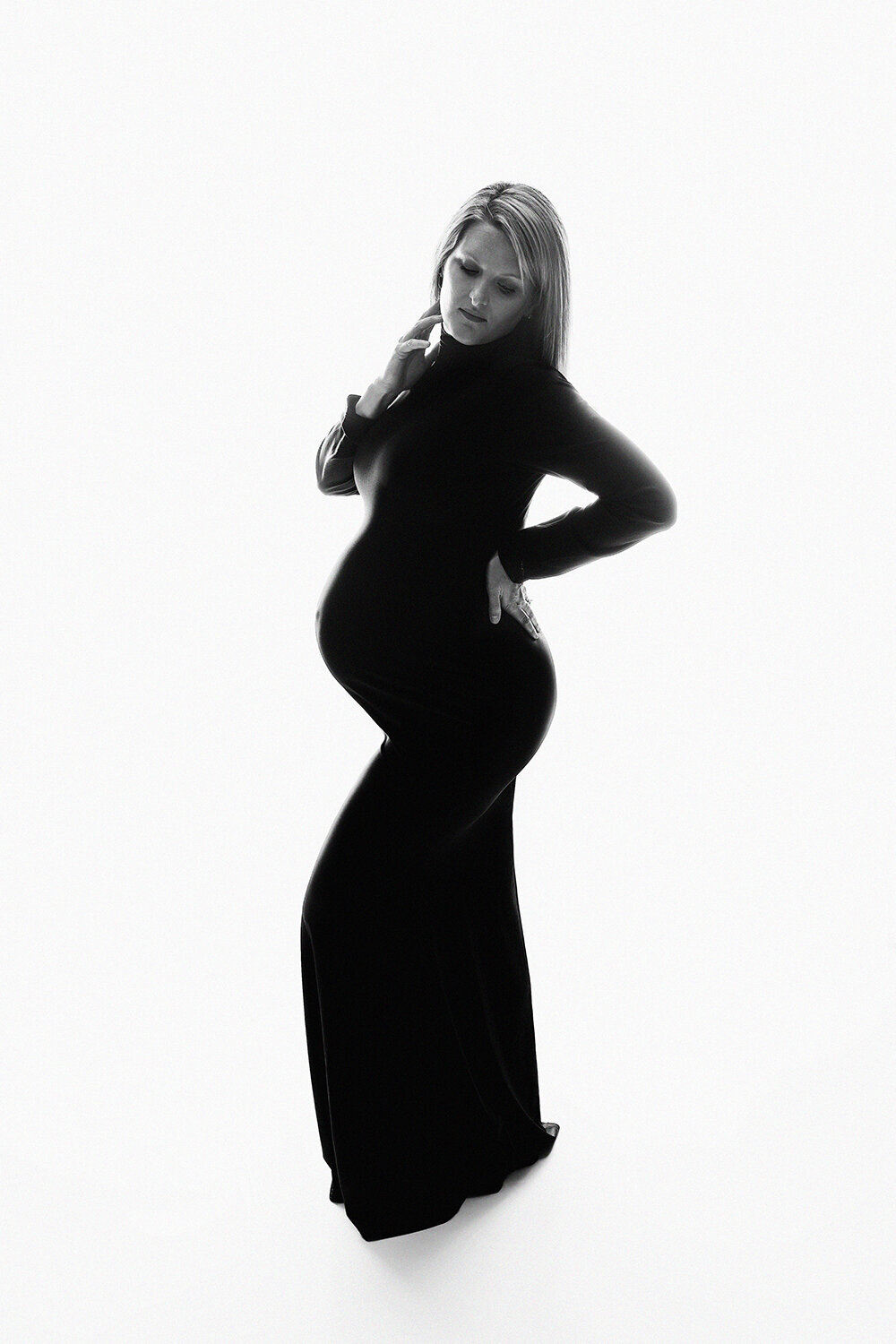 B&W photo of a woman stands in a studio with a hand on her back in a black maternity gown