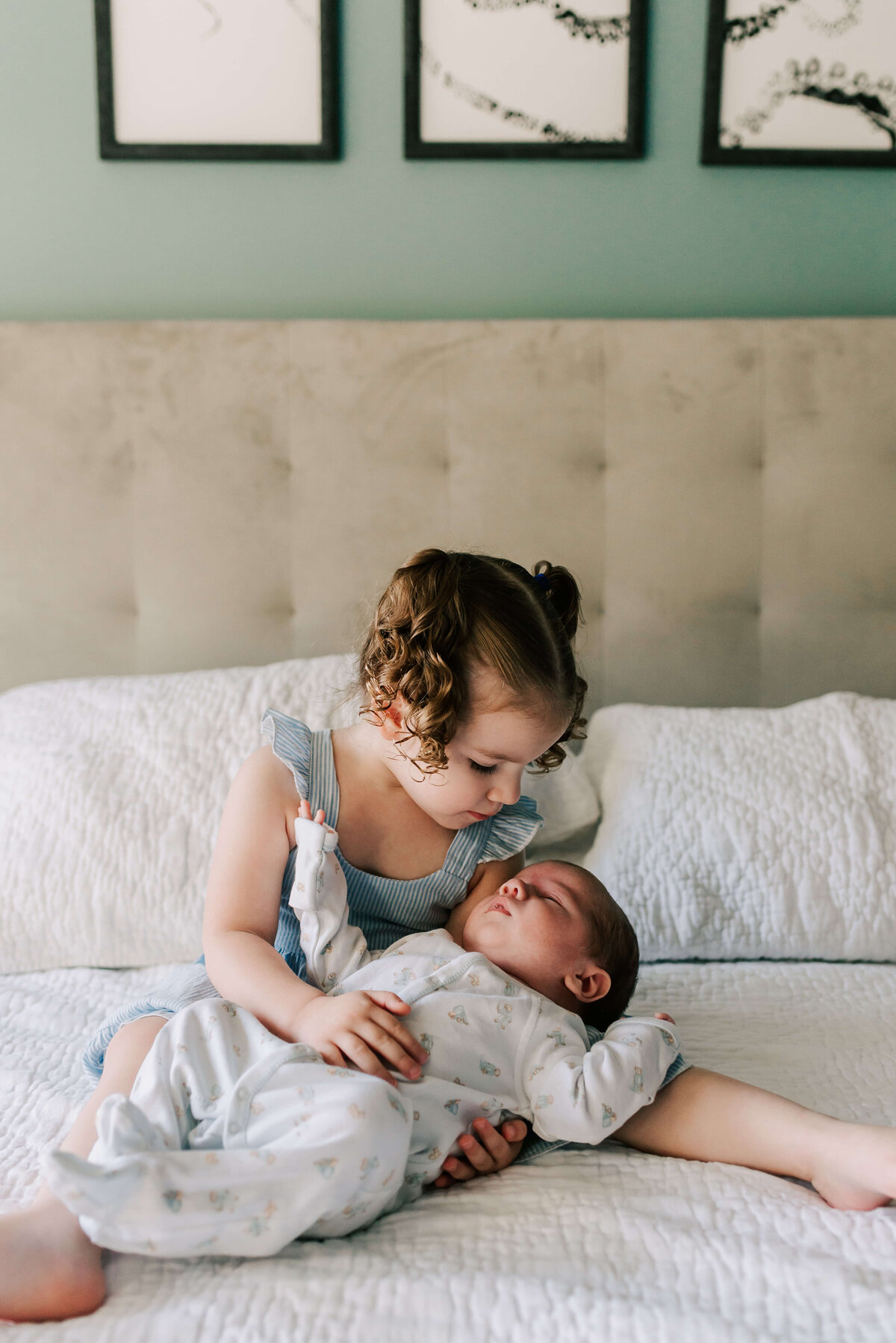 A new big sister sitting on a bed holding her baby brother, taken by Denise Van Photography