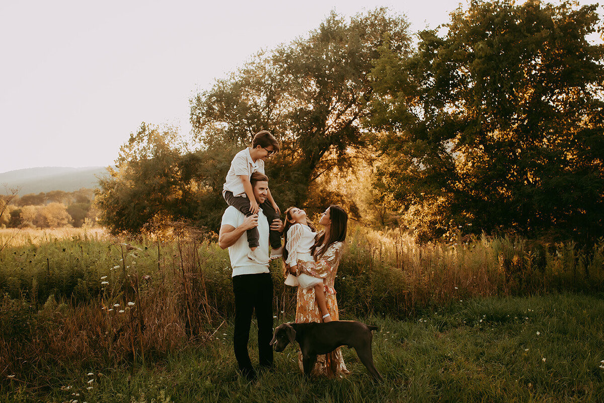 a family of four standing in a field with their dog looking at each other during golden hour