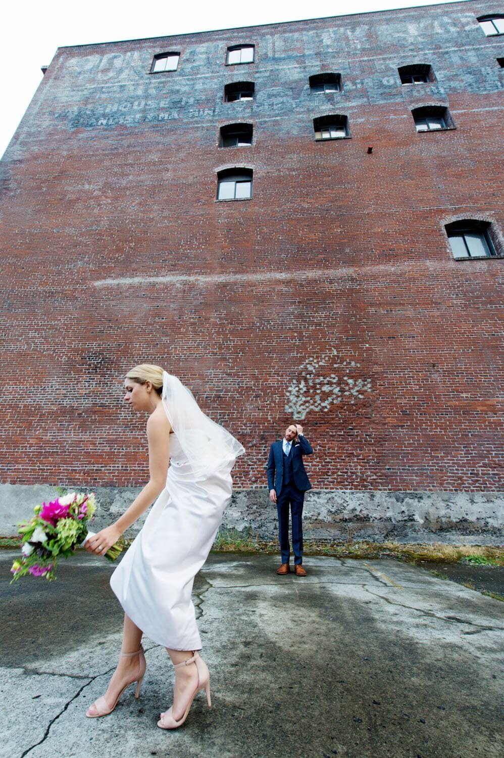 a bride puts her bouquet down as a groom waits for her in front of a tall brick building in nw portland