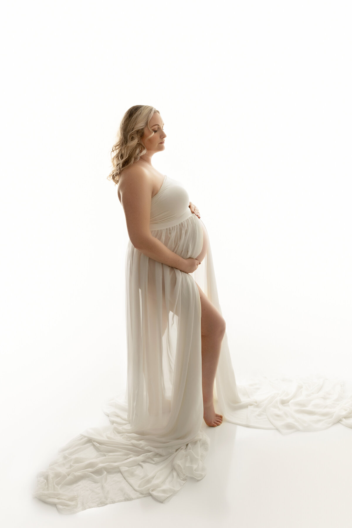 A pregnant woman in a sheer maternity gown stands in a studio holding her bump