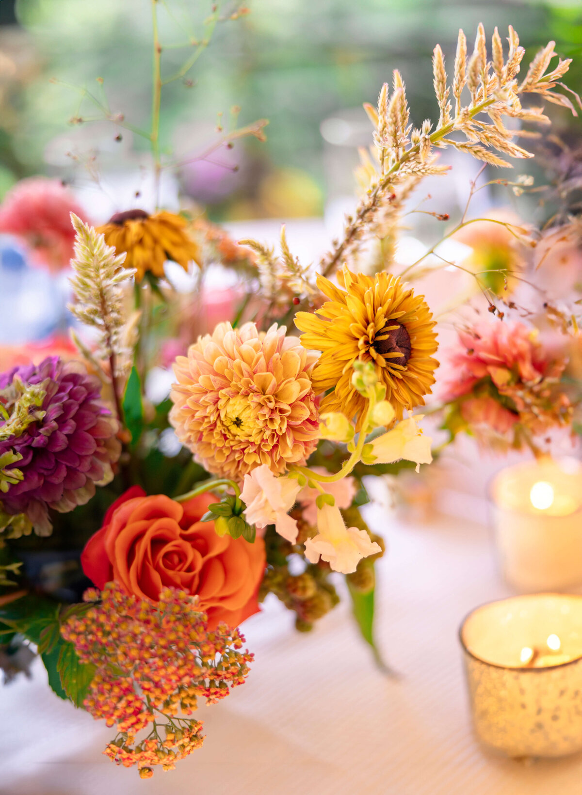 A vibrant floral arrangement featuring orange roses, pink and purple dahlias, and yellow sunflowers, surrounded by greenery and delicate wildflowers. Soft candlelight creates a warm and inviting atmosphere.