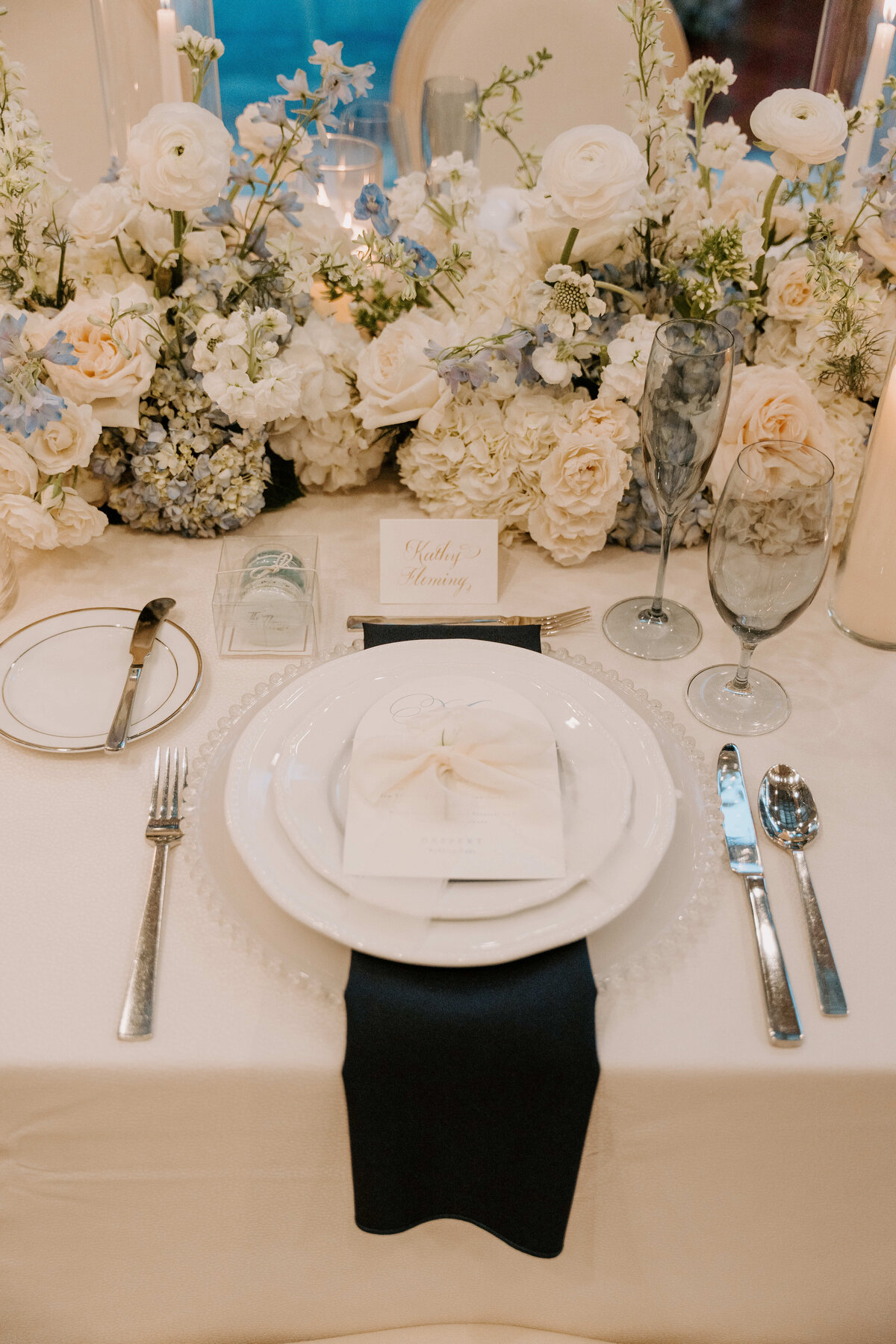 Elegant wedding table setting with white flowers and greenery centerpiece. White plates on black napkin, silver cutlery, and glassware convey sophistication.