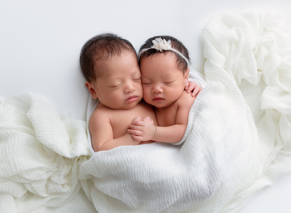 Twin brother and sister pose for Brooklyn, NY newborn photoshoot. Brother is cuddling his sister with his arm resting on her back. Sister has her hand resting on brother's belly. Both babies are sleeping and draped in white muslin frabric.