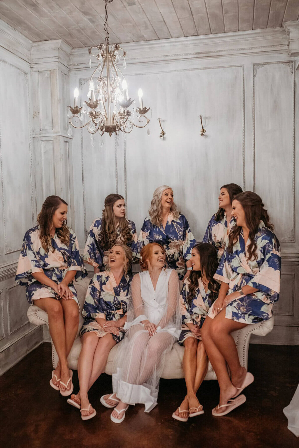 Photo of women and white and navy robes sitting on a couch laughing together with a chandelier overhead