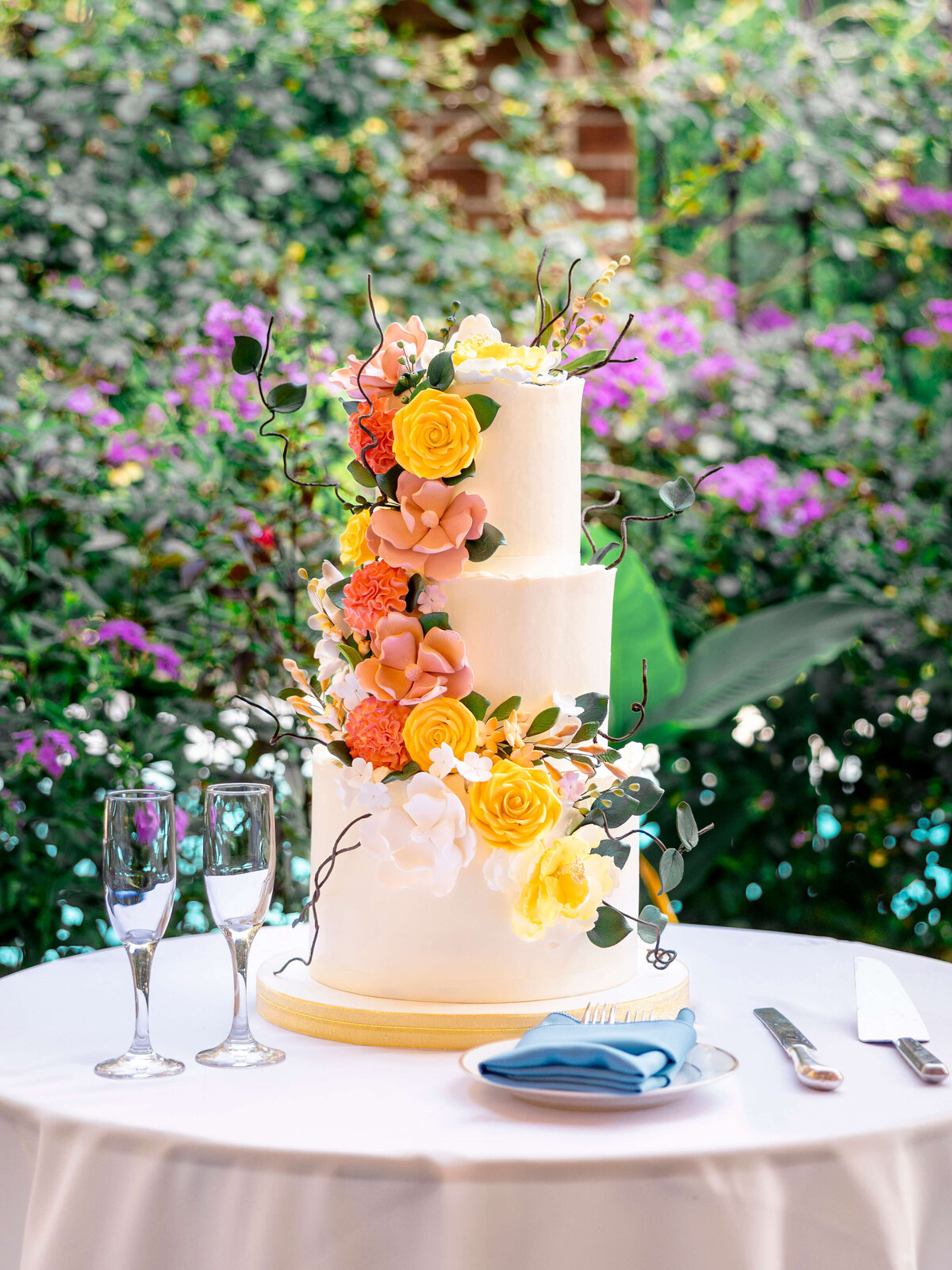 A three-tiered white cake adorned with vibrant flowers in yellow, pink, and orange sits on a round table. Two empty champagne flutes, a fork, knife, and blue napkin are arranged beside it. The background features lush greenery and purple blossoms.