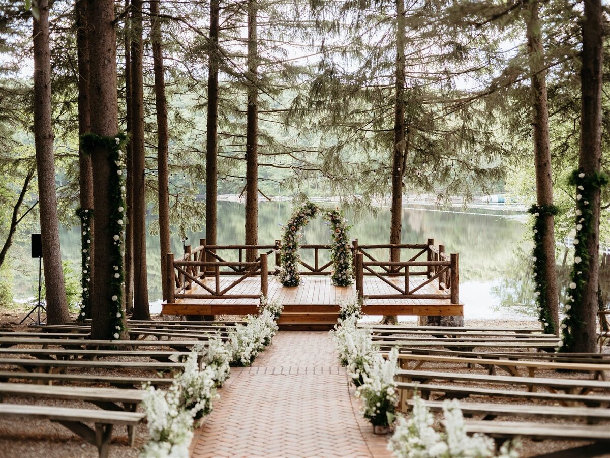 Stunning floral arbor by Canvas Weddings sits on wooden dock overlooking lake and trees at Cedar Lakes Estate wedding venue in Catskills; aisle lined with white florals; under pine trees; long wooden benches for guests