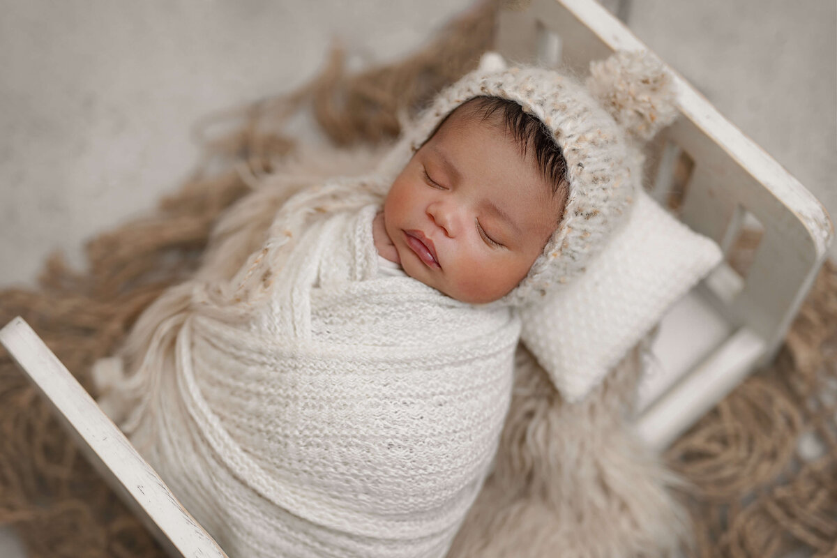 newborn swaddled in cream with bonnet in white wooden bed