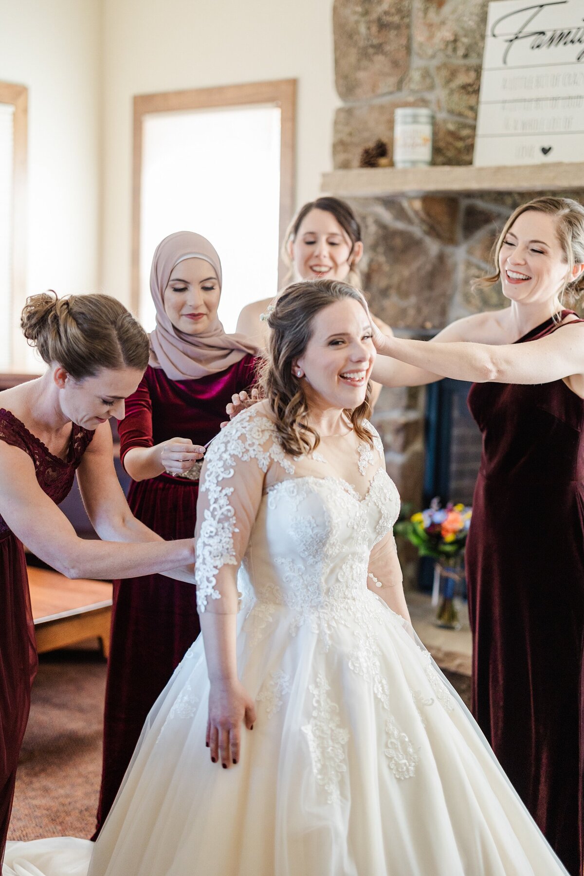 A bride standing and laughing as her bridesmaids help lace up her wedding dress before her ceremony at the YMCA of the Rockies in Estes Park, Colorado. The bride is wearing a long, flowing white dress with floral sleeves. The bridesmaids are all smiling and helping out while wearing dark burgundy dresses.