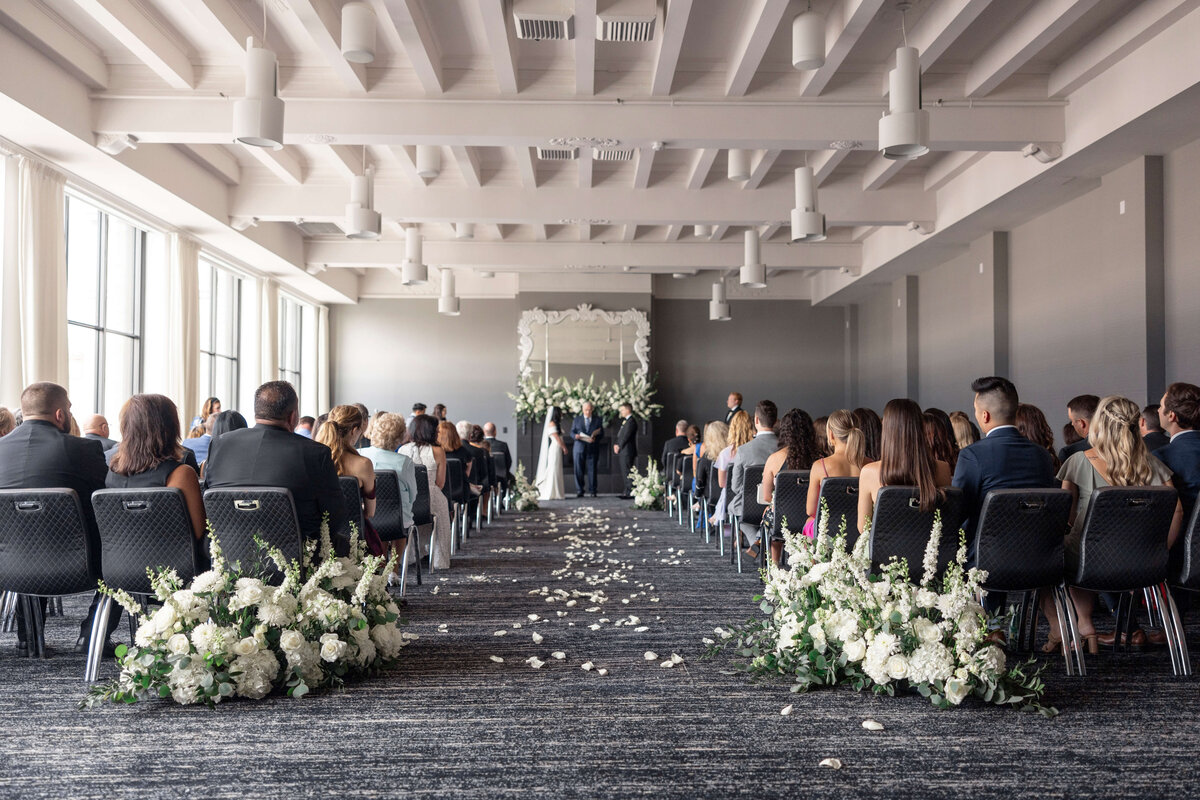 A wedding ceremony in a bright, modern venue with white flowers lining the aisle. Guests are seated on either side, facing the altar where the couple stands with the officiant. Large windows on the left let in natural light.
