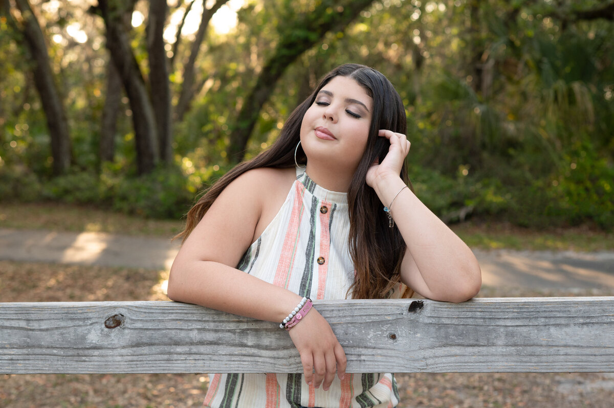 High school senior girl with eyes closed smiling leaning on a wooden fence.