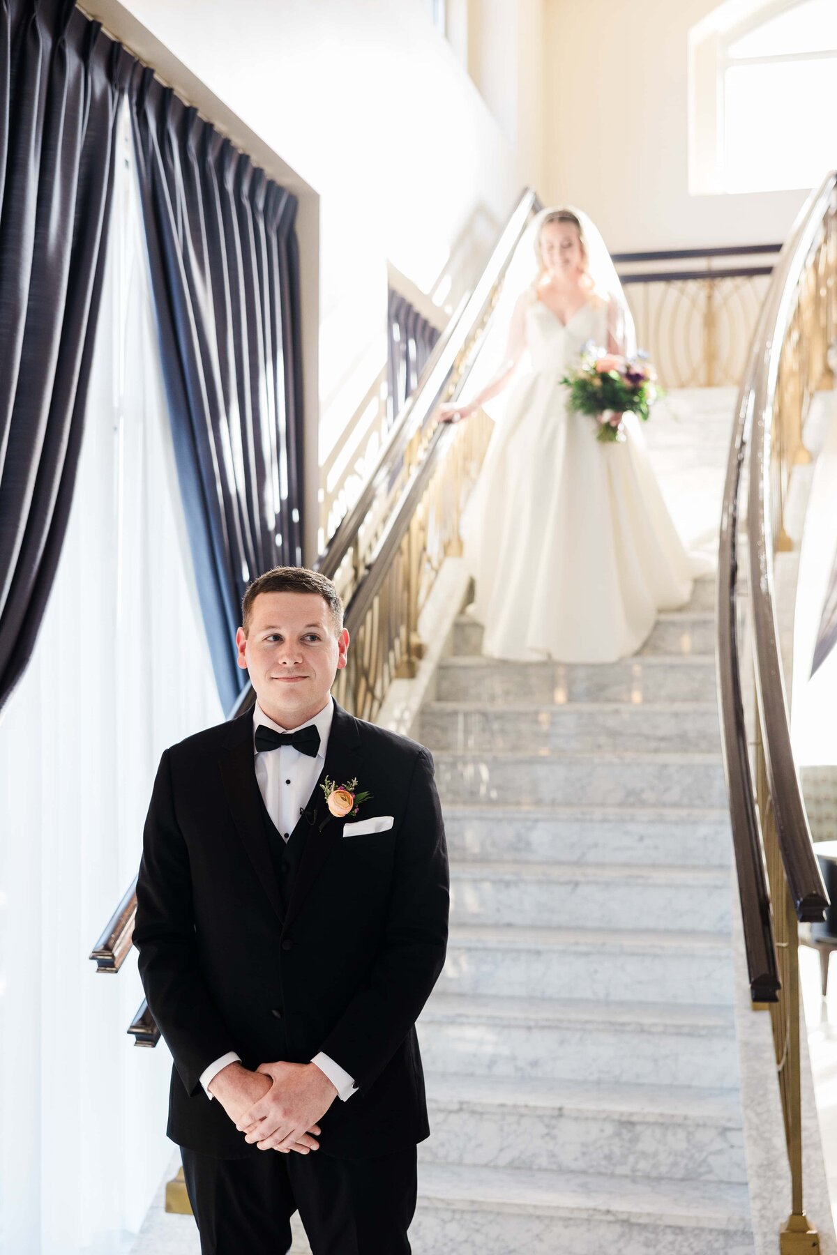 A bride in a white dress descends a staircase behind a groom in a black tuxedo waiting at the bottom, in an elegant park farm winery setting.