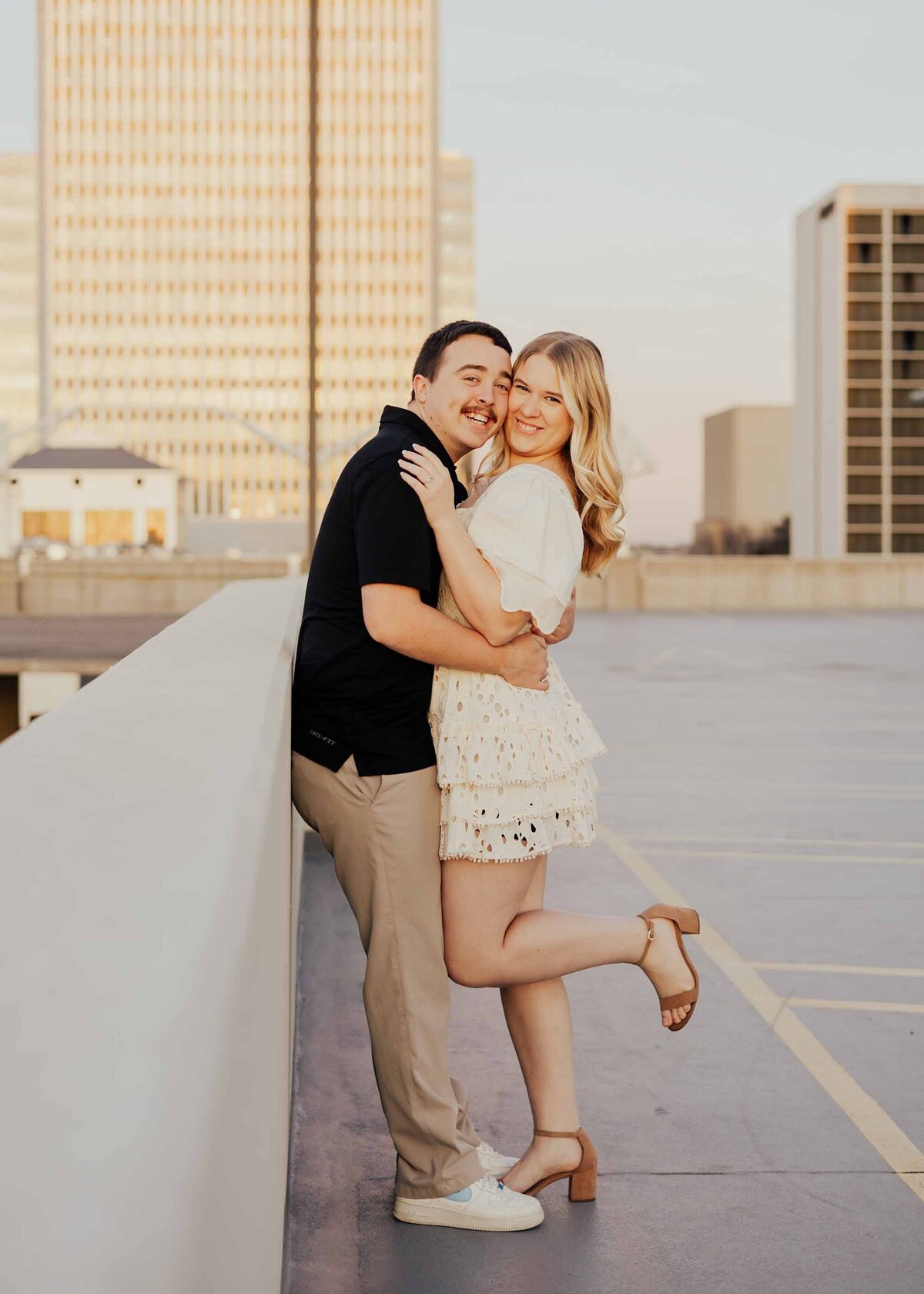 Maddie Rae Photography couple standing and leaning on the wall of a parking garage. face to face looking and smiling at the camera. he has her pulled in by the waist and she has one leg lifted and bent