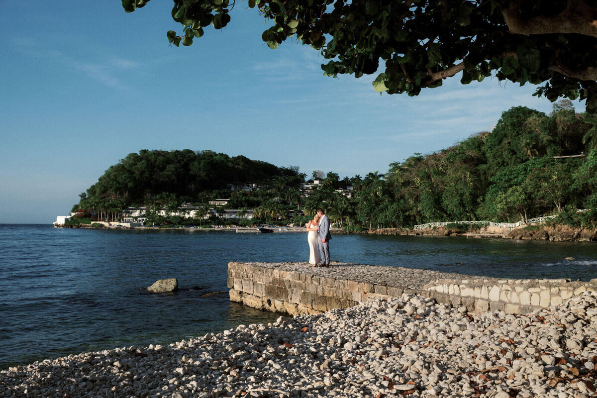 The engaged couple is standing on a stone dock in Round Hill Hotel and Villas, Jamaica. Ocean and mountains in the background