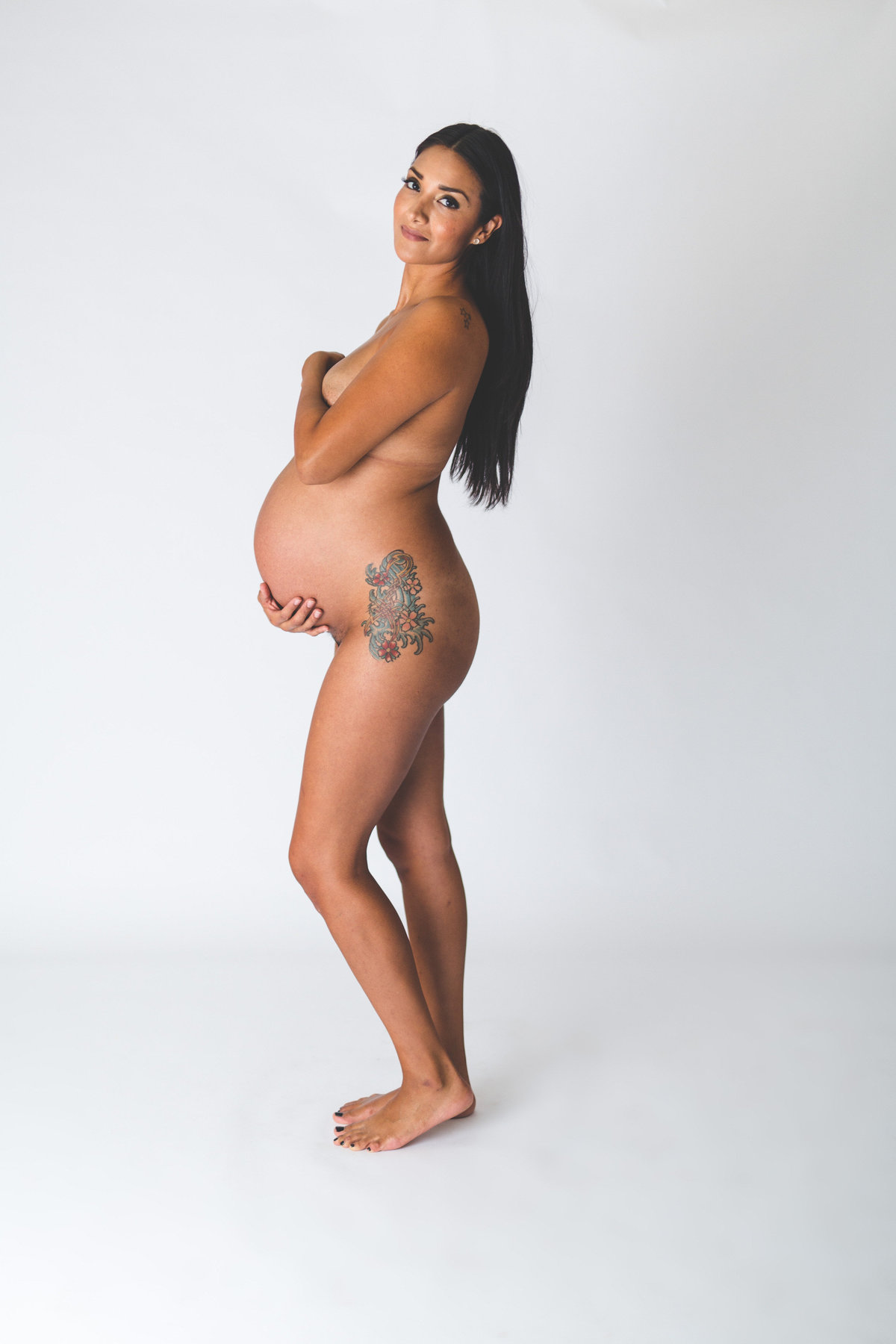 Maternity picture of woman covering herself with her hands and posing in front of white backdrop.