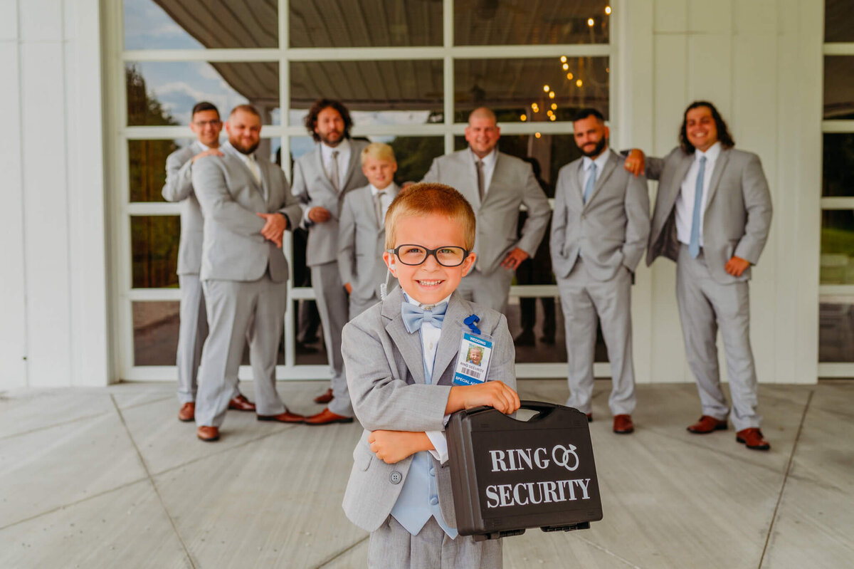 A photo of a ringbearer holding a ring security box with the groomsmen posed in the background