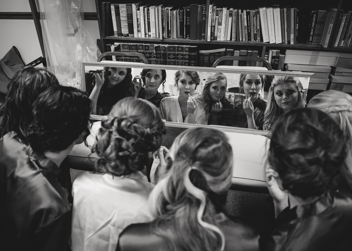 black and white image of bride and bridesmaids looking in mirror putting on makeup