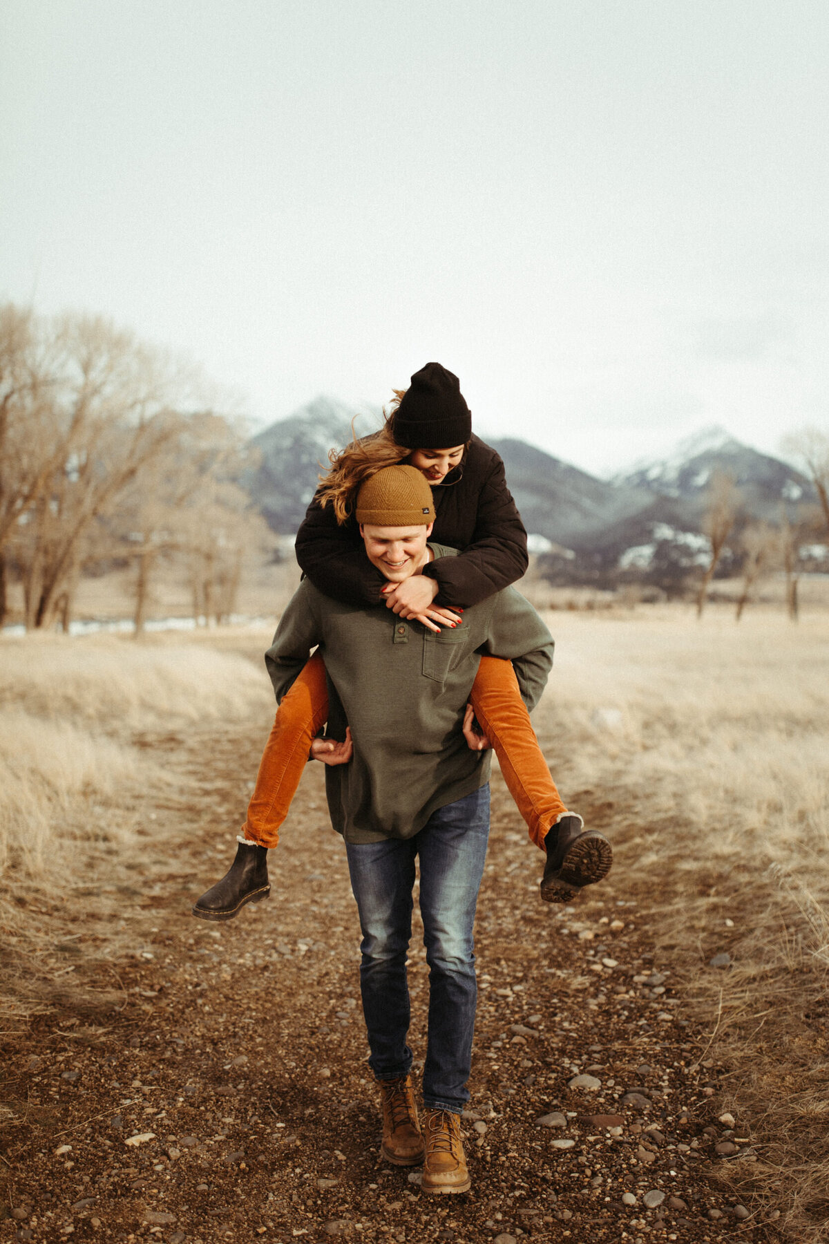 Guy giving his girl a piggy back ride down a trail with mountains in the background on a cold winter day
