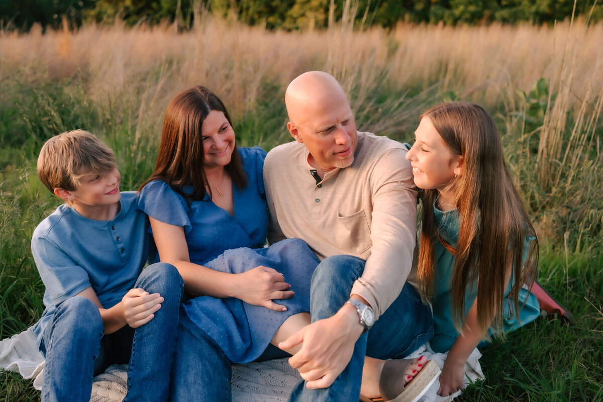 A family of four are sitting on a blanket in a field while looking and laughing at each other just having fun with their portrait session.