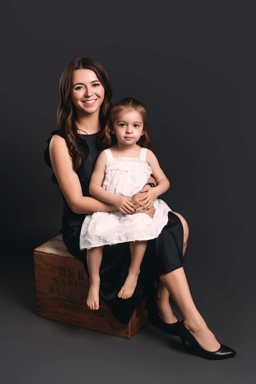 A mother in a black dress and heels sits on a vintage box in front of a gray-toned backdrop holding her young barefoot daughter in a white dress, captured by a photographer in Bemidji, Minnesota.