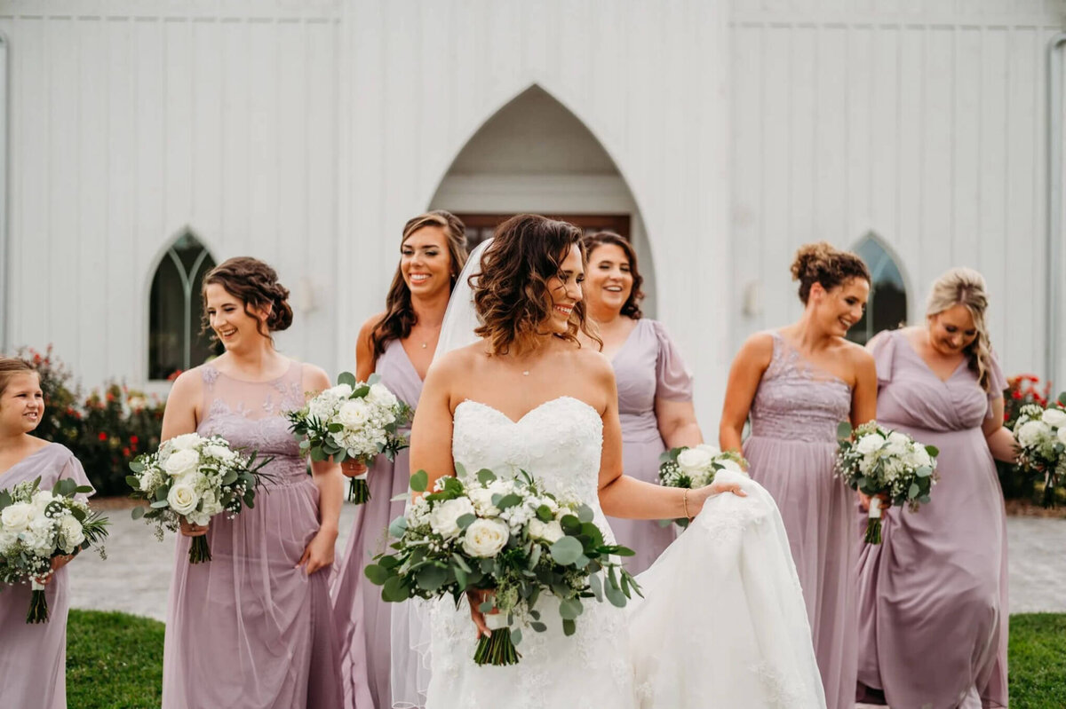 Photo of a bride caring the train of her dress and smiling while her bridesmaids walk in the background