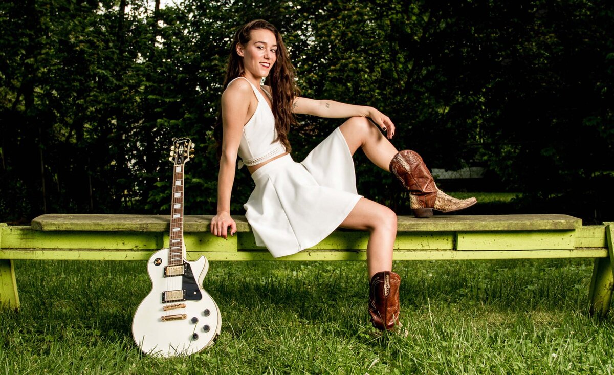 Female musician photo Leanne Pearson wearing white dress red cowboy boots sitting against green bench beside white electric guitar upon green grass green trees background