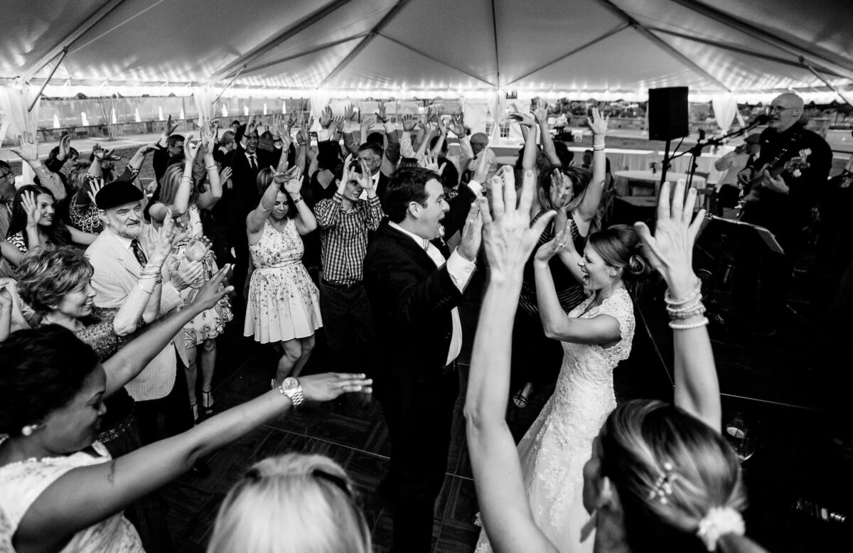 A black and white photograph captures the jubilant atmosphere of a wedding reception, with guests raising their hands in the air, dancing around the newlyweds.