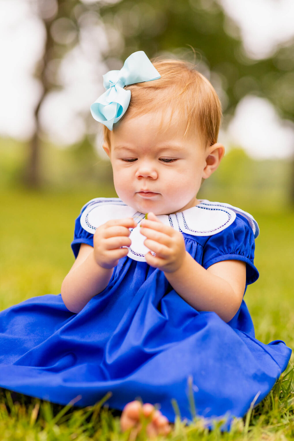 little girl in a blue dress sitting in the grass holding a flower