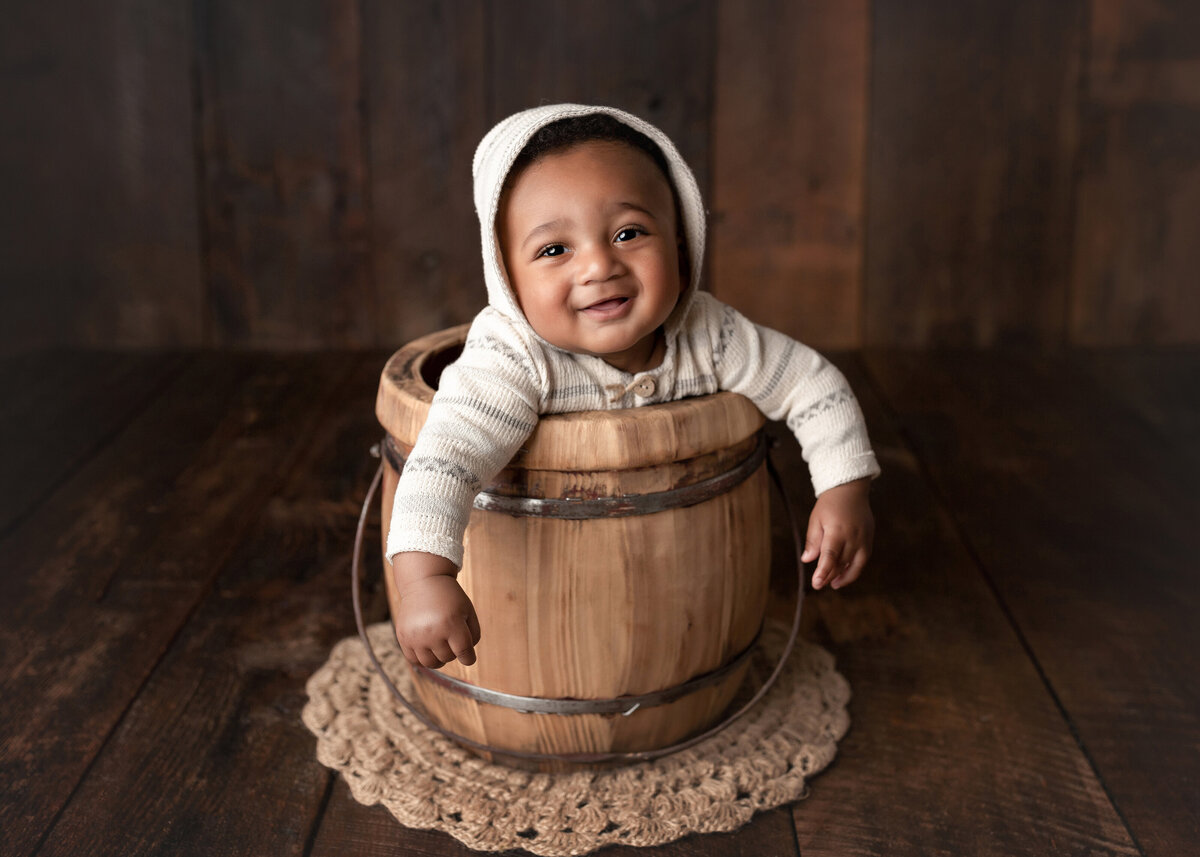 Baby boy 9-month milestone photos. Baby is sitting in a bucket with his arms draped over the edge. Baby is in a knit sweater and hood smiling at the camera. Bucket is on a barn wood floor and matching barn wood backdrop.