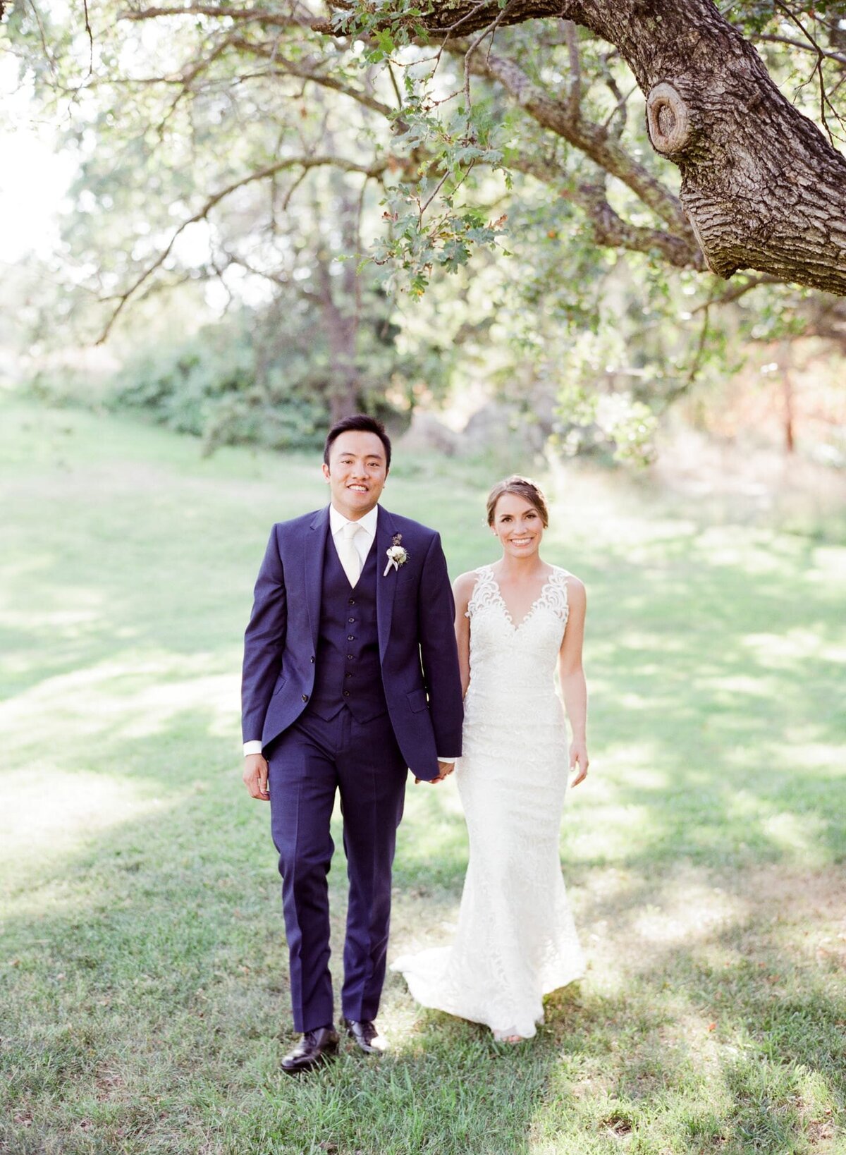 After an impressive farm wedding at a farm, newly-wed couple hold hands and walk towards the camera beneath a soothing tree shade.