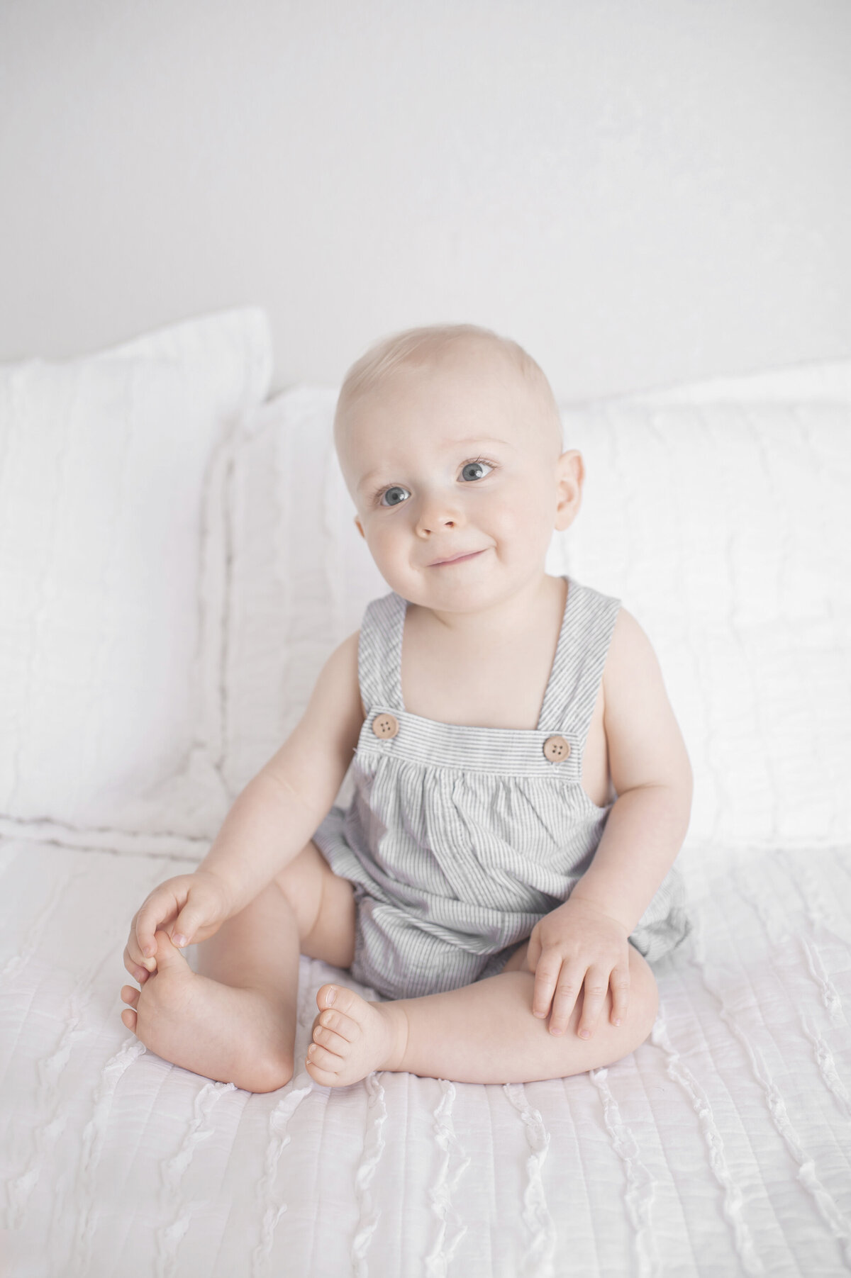 one year old baby sitting on white bed holding foot smiling
