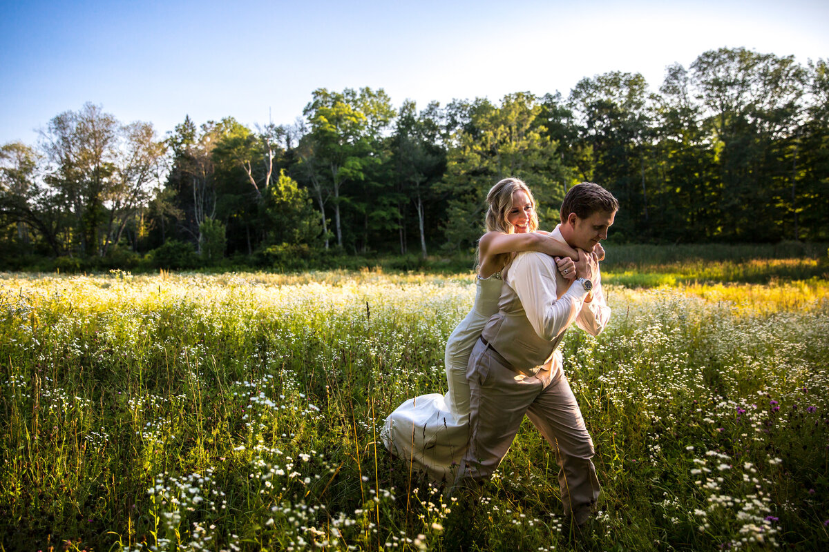 A groom carrying a bride on his back through a field.