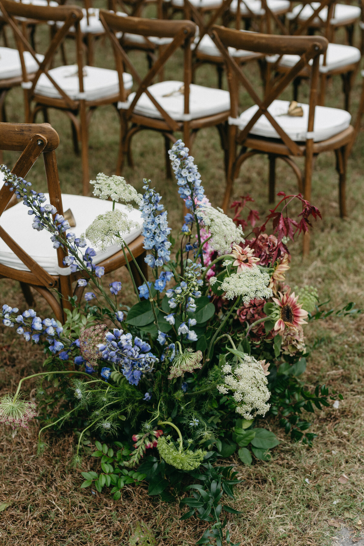 Ceremony aisle floral installation featuring blue delphinium, queen anne’s lace, dahlias, and lush greenery beside wooden cross-back chairs at an outdoor garden wedding.