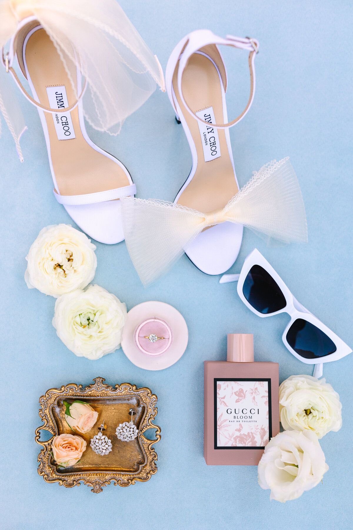 flat lay image of bride's shoes, sunglasses, perfume, jewelry, and flowers on blue background.