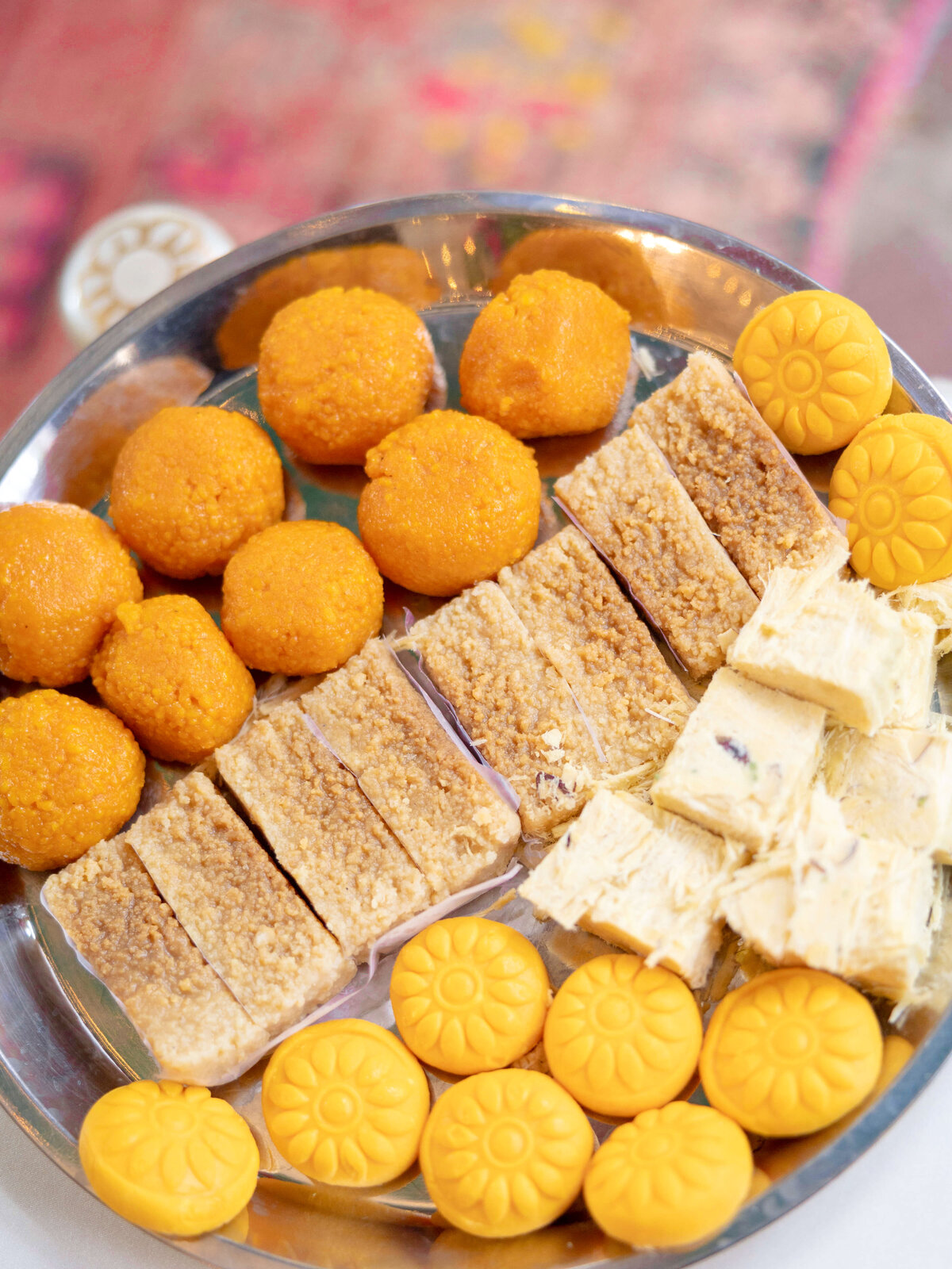A plate with an assortment of Indian sweets, including round orange ladoos, rectangular beige barfis, and yellow modaks arranged neatly. The sweets are set on a metallic tray.