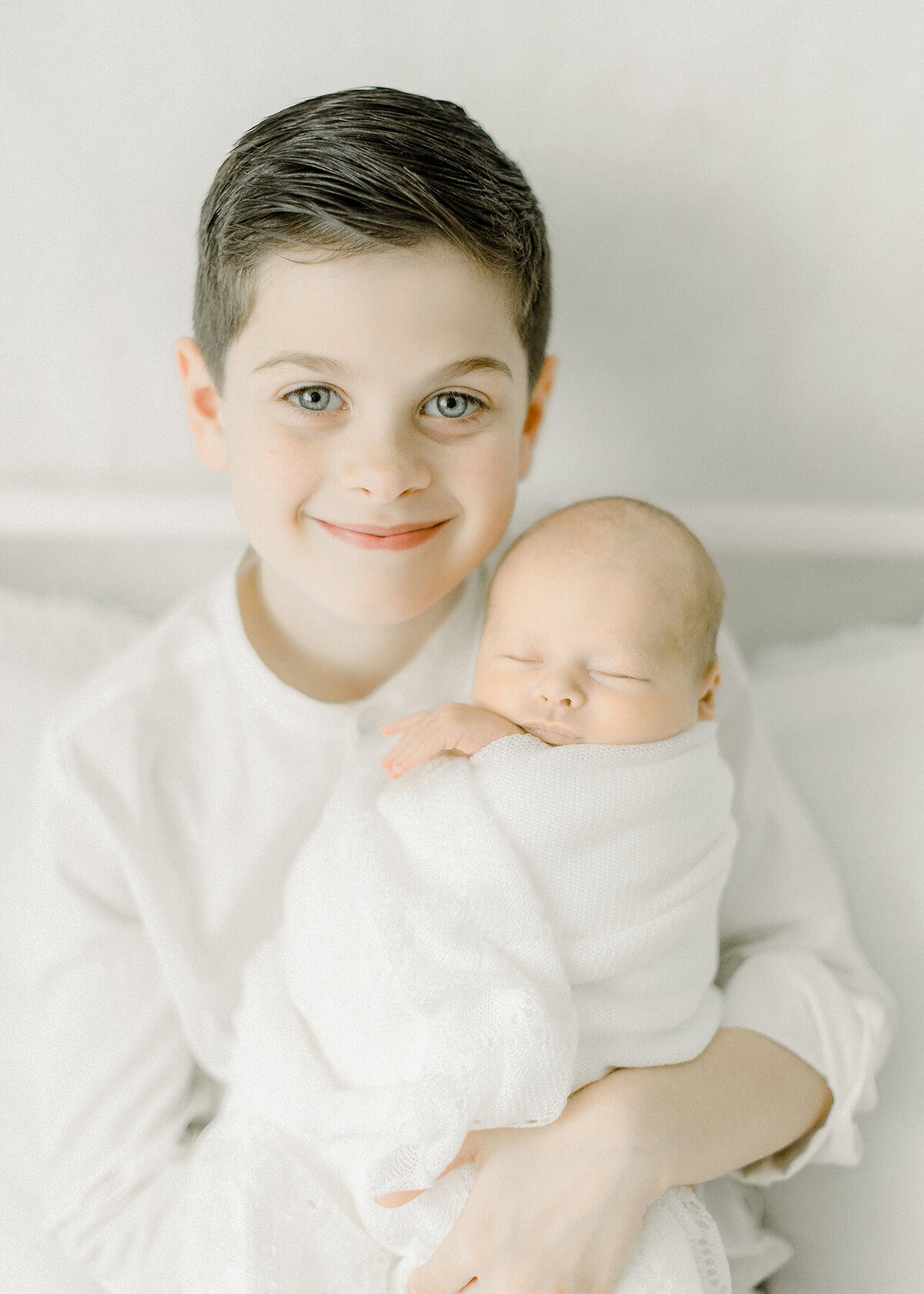 Proud big brother holding his newborn baby brother close as he looks at the camera and smiles while sitting on a bed in a Dallas photography studio.