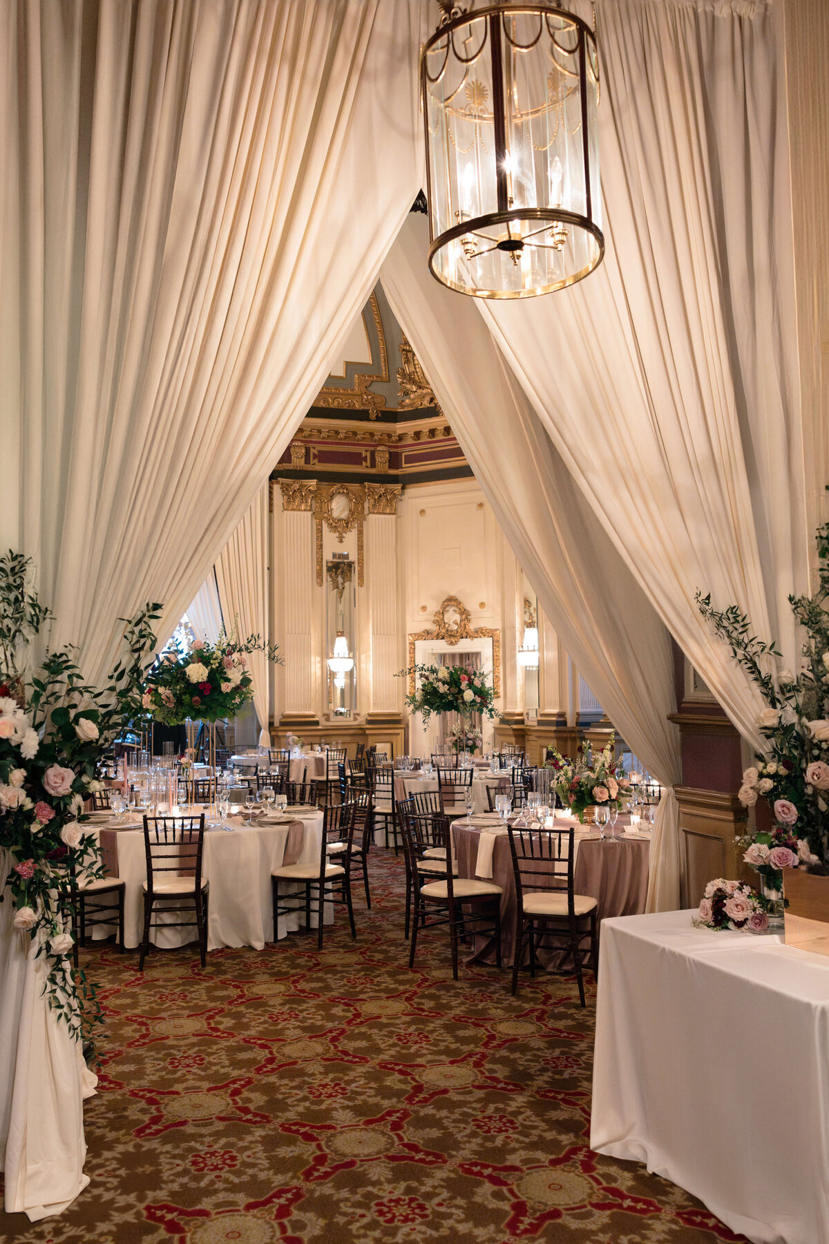 Elegant wedding reception with round tables set for dining in an opulent hall. Tall floral centerpieces and candlelit chandeliers enhance the ambiance. White drapes frame the entrance, leading into the ornate, classic interior.