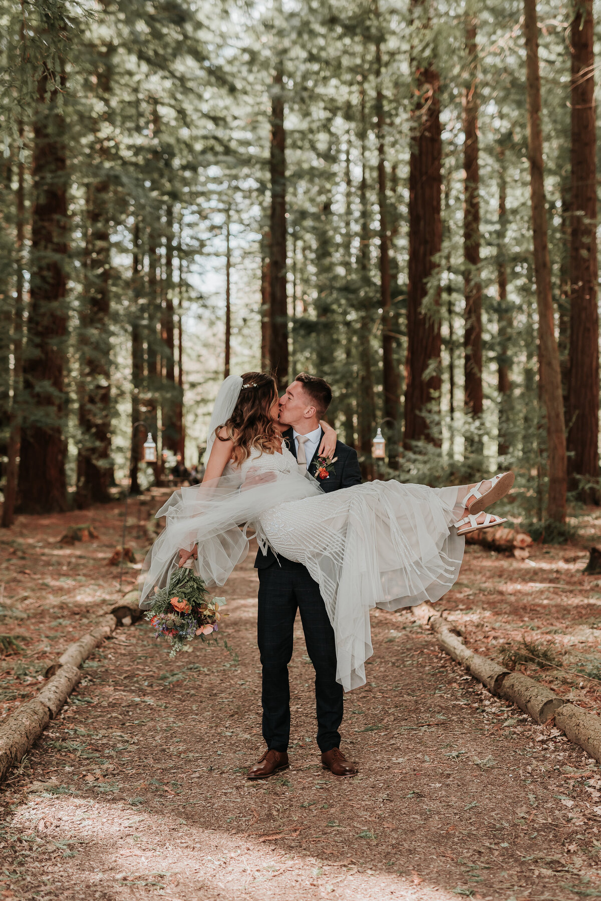 Groom carries his Bride along a woodland pathway at their intimate woodland wedding at Two Woods Estate